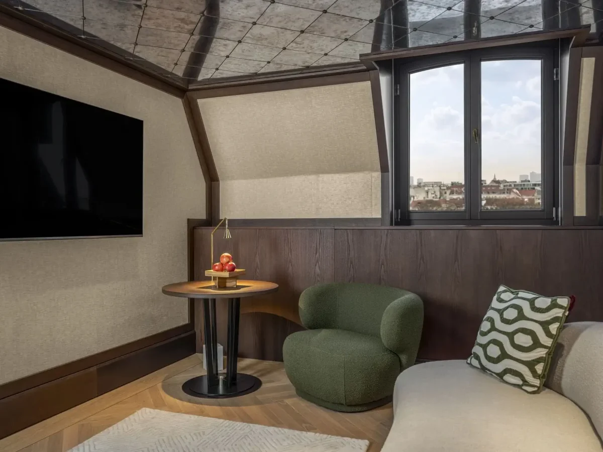 A modern Almanac Park Suite features a wall-mounted TV, a round table with a lamp and apples, a green chair, a beige sofa with a patterned pillow, and a window offering a city view.