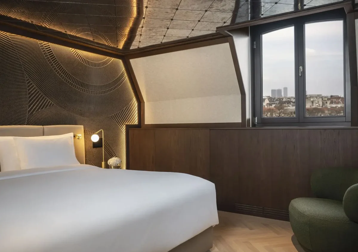 Experience the Almanac Park Suite: a modern hotel room featuring a large bed, textured accent wall, wood paneling, a city-view window, and a stylish green armchair.