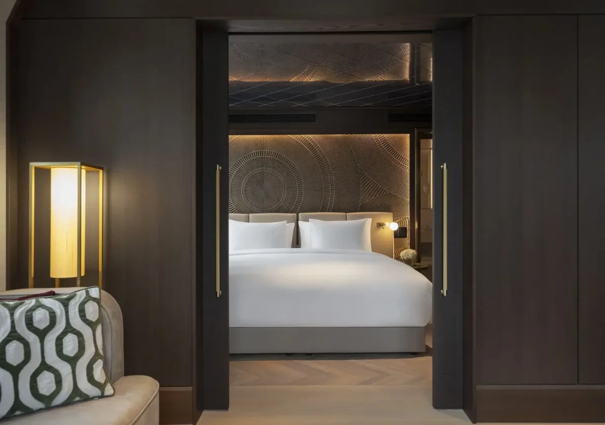 A modern Palais Suite bedroom with a large bed, white linens, patterned headboard, sliding dark wood doors, and ambient lighting. A sofa with a geometric cushion is partially visible.