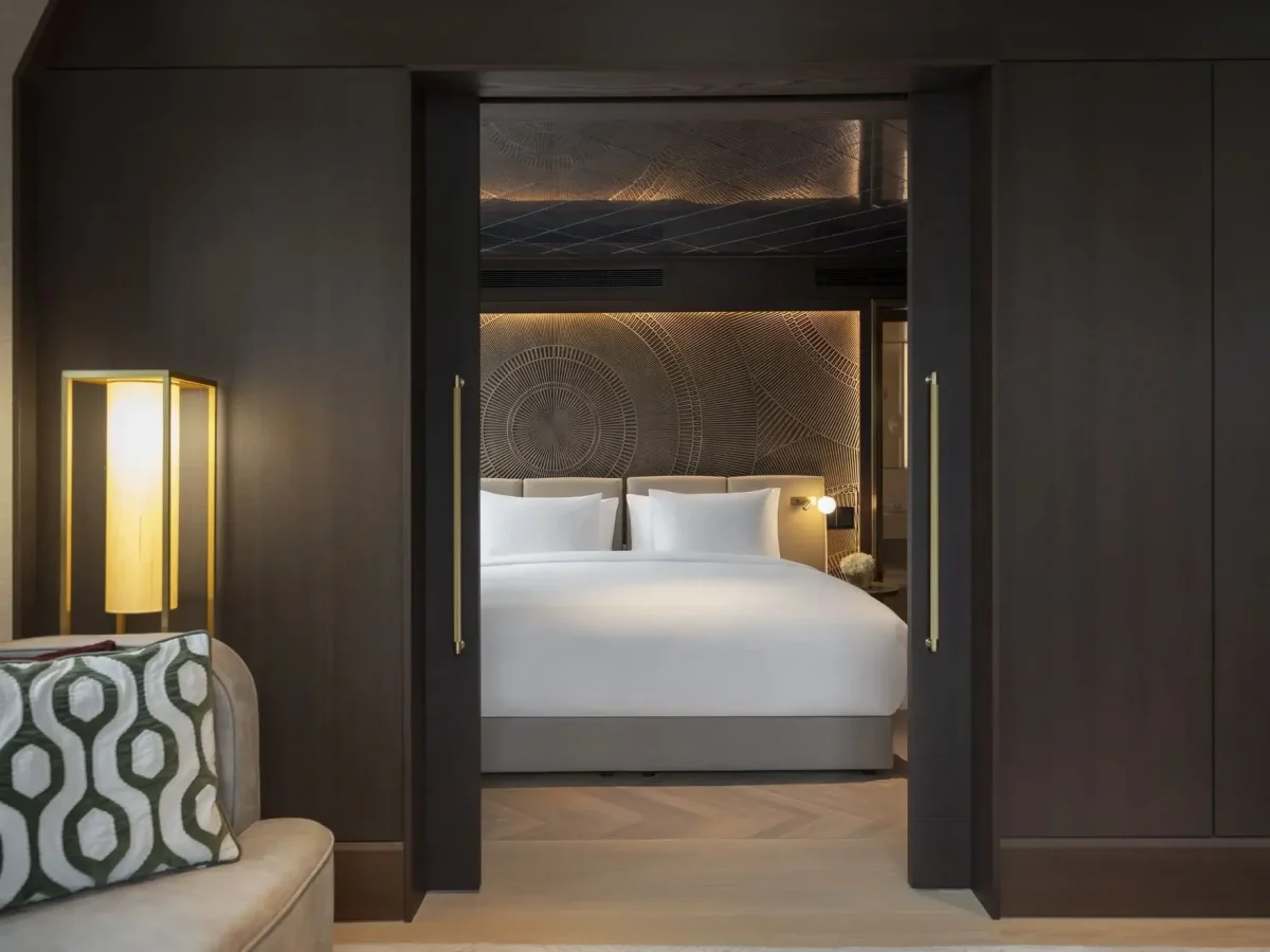 A modern Palais Suite bedroom with a large bed, white linens, patterned headboard, sliding dark wood doors, and ambient lighting. A sofa with a geometric cushion is partially visible.