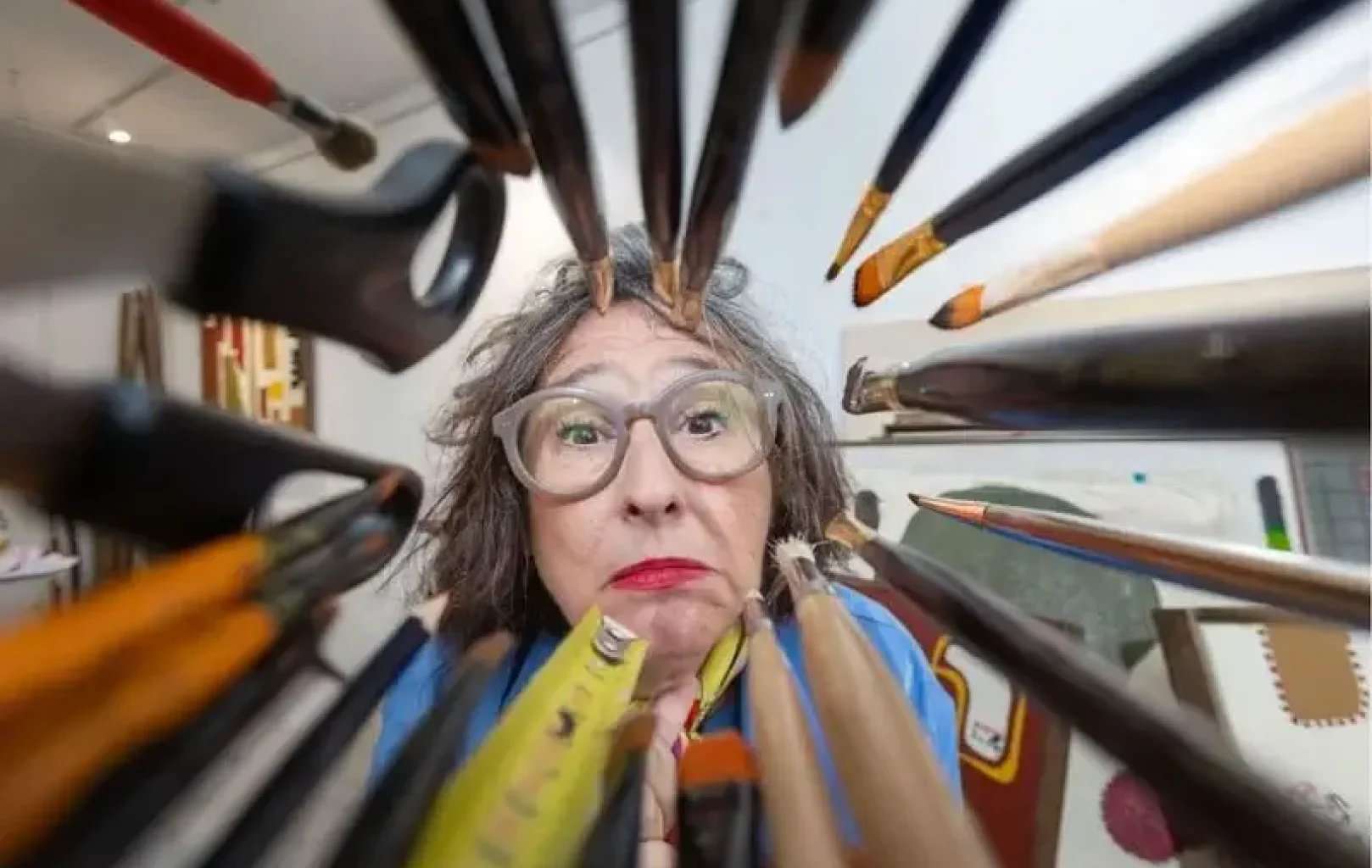 Una persona amb els cabells grisos i ulleres mira a través d'un grup de pinzells i llapis d'art, amb la cara emmarcada per les eines en primer pla.