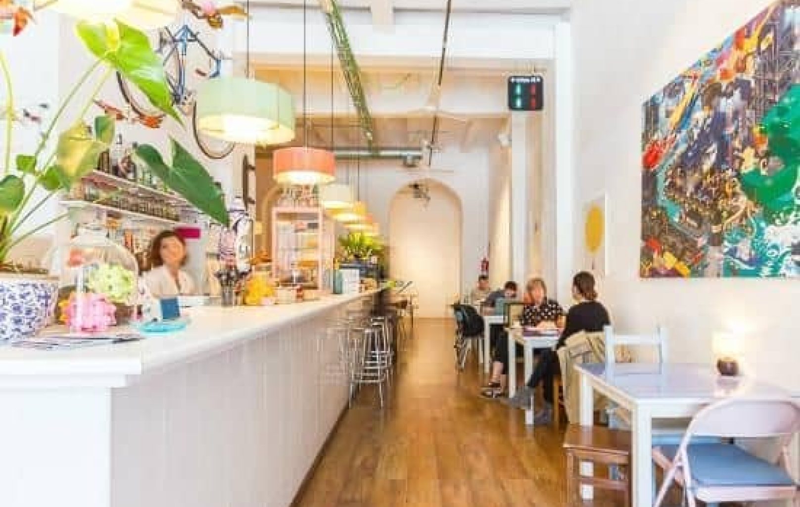 Bright, modern café with a long white counter, colorful hanging lights, and several people seated at tables along the wooden floor.