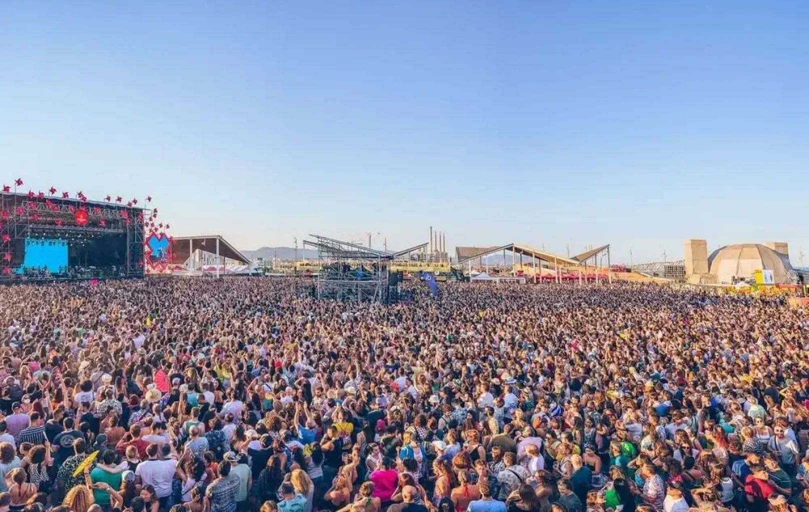 Una gran multitud se reúne al aire libre frente a un escenario en un festival de música bajo un cielo azul despejado, con carpas y estructuras al fondo, capturando la vibrante energía de las celebraciones de Junio y Julio.