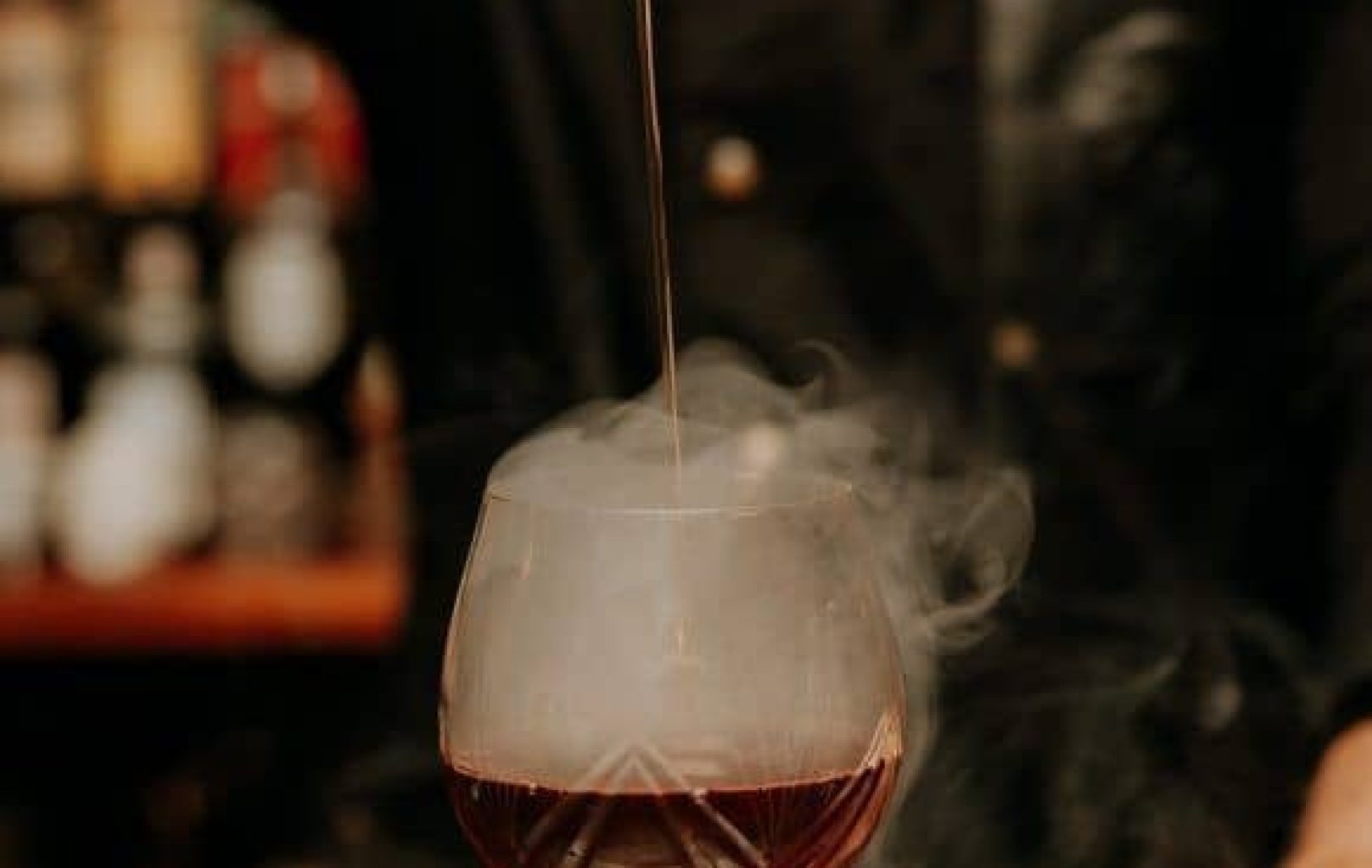 A bartender pours a liquid into a glass of dark cocktail emitting smoke on a bar counter, with shelves of Dux Gins and other bottles blurred in the background.