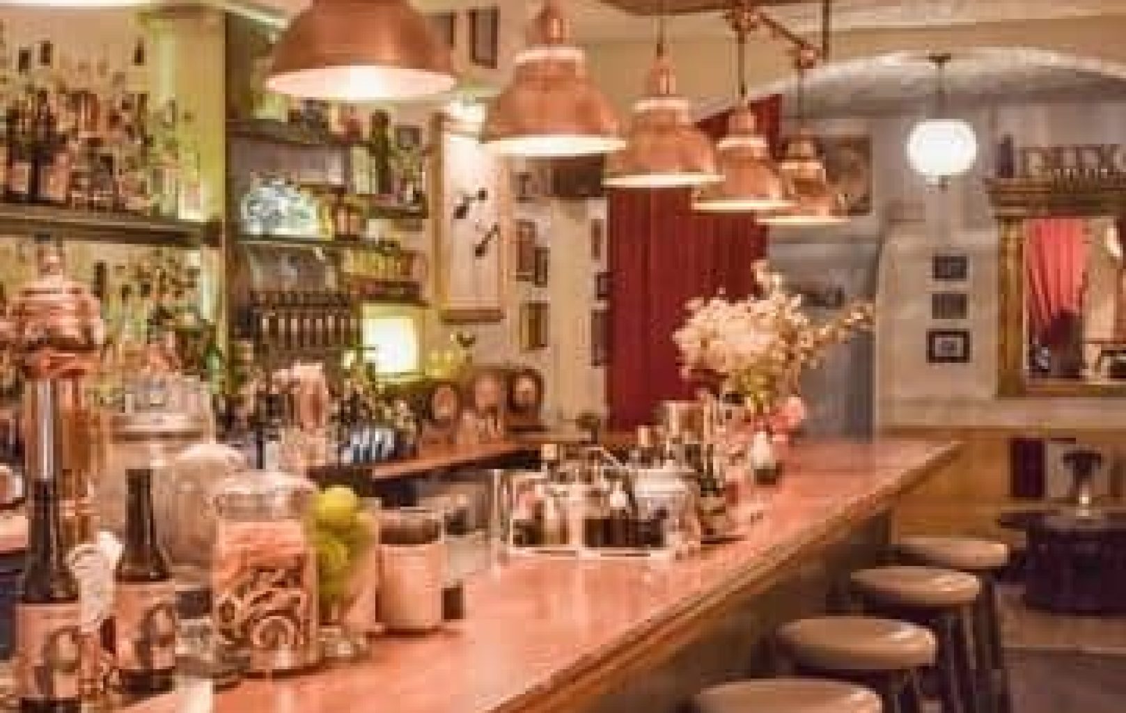 A cozy bar with a wooden counter, high stools, hanging pendant lights, and shelves stocked with bottles and glassware serving signature cocktails made with Dux Gins.