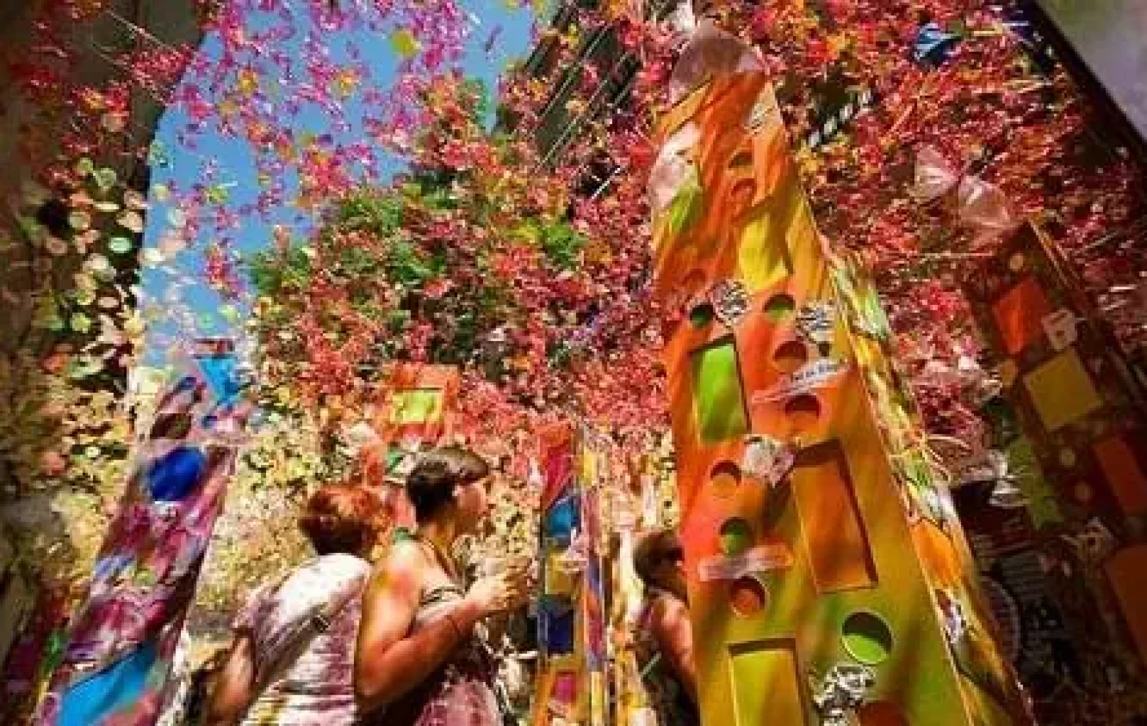 La gente pasea por una calle decorada con coloridas estructuras altas y adornos colgantes bajo un cielo azul brillante, celebrando las vibrantes tradiciones de Junio y Julio.