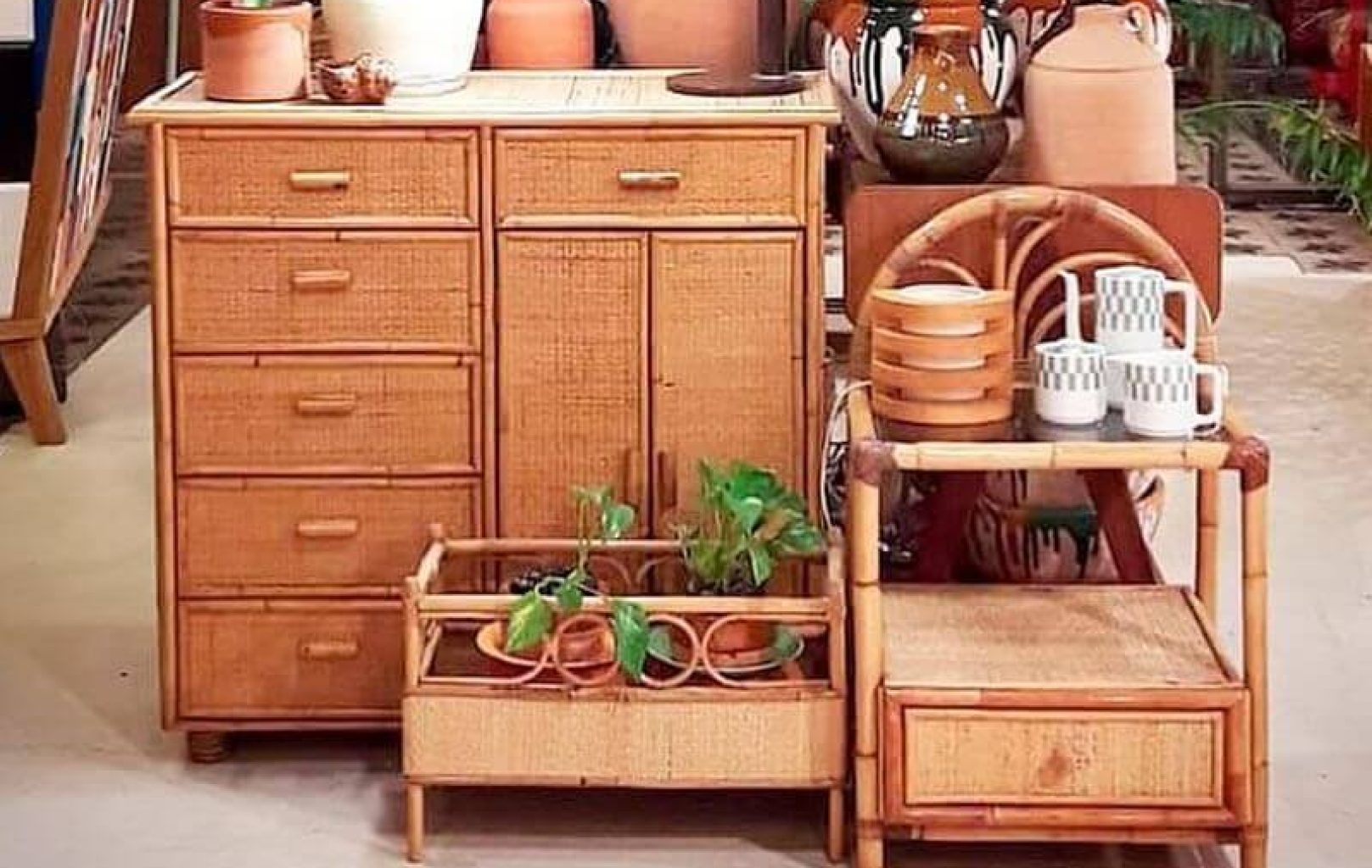 Una cómoda de madera, un soporte para plantas con macetas verdes y una silla de bambú que sostiene tazas y platos se exponen en una tienda con otros objetos de alfarería y cerámica al fondo.
