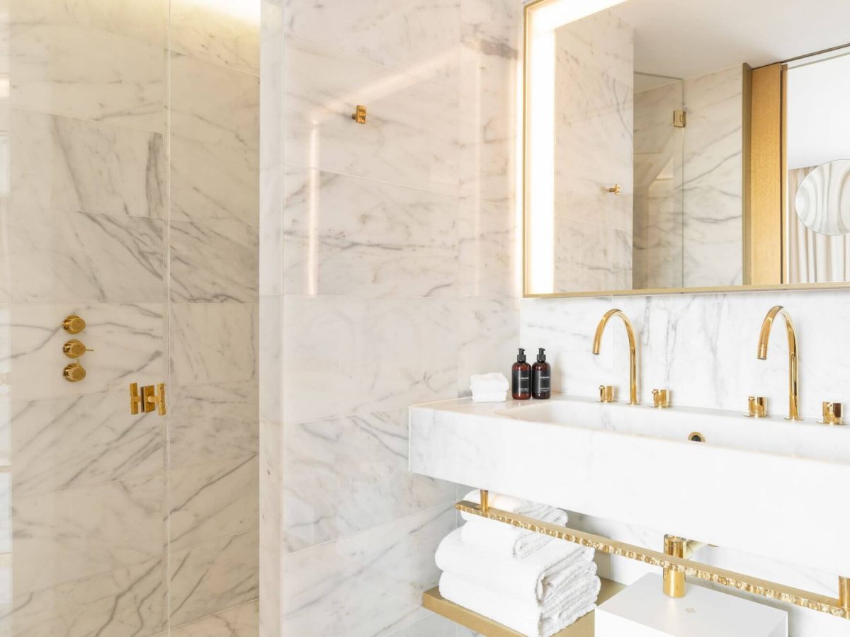 Moderno cuarto de baño con paredes de mármol blanco, lavabo doble, espejo grande y accesorios dorados, que evocan un ambiente de Gallery Room. Debajo del lavabo se exponen toallas blancas perfectamente dobladas en estantes abiertos.