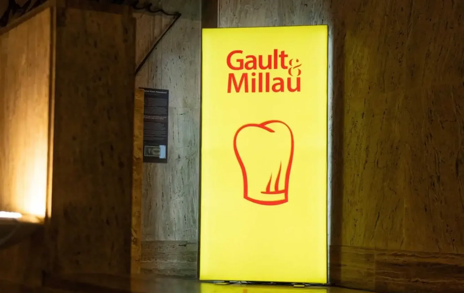Zářivě žlutá cedule s červeným nápisem "Gault&Millau 2025" a grafikou kuchařské čepice na mramorové stěně v interiéru Restaurace Alcron.