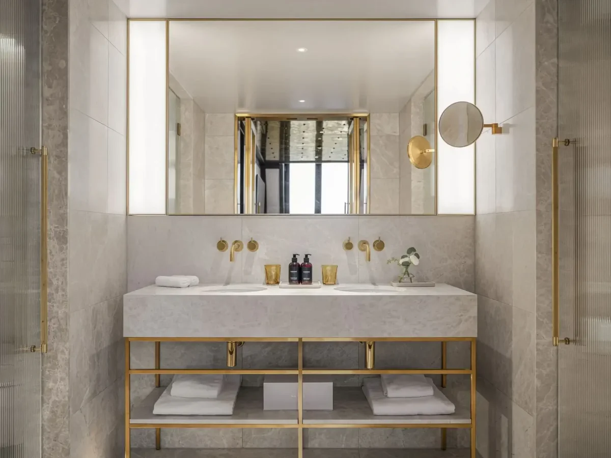 Modern Duplex Park Suite bathroom with double sinks, marble countertop, gold fixtures, large mirror, toiletries, rolled towels, and shelves below holding additional towels.