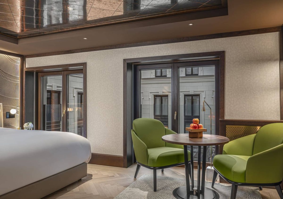 An Almanac Room features a large bed, two green armchairs around a small round table with fruit, and floor-to-ceiling windows overlooking another building.