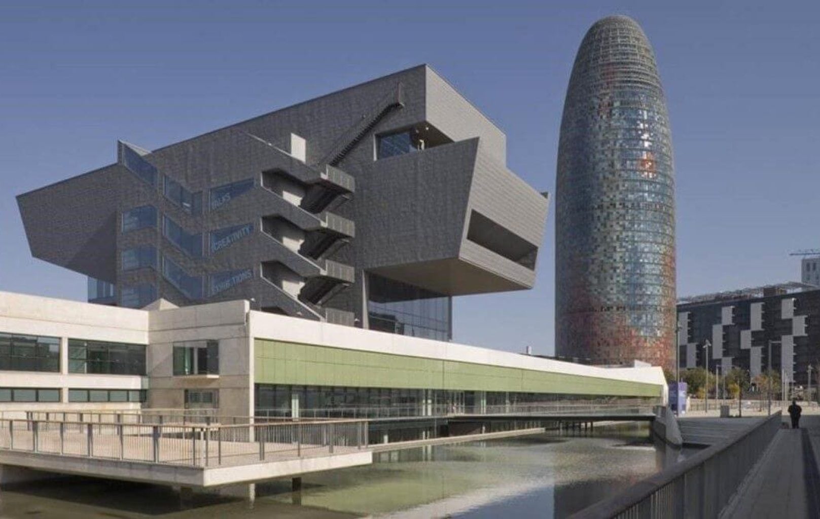 Modern architectural buildings with geometric shapes beside a reflective pool, featuring the cylindrical Torre Glòries in Barcelona under a clear sky, evoke the spirit of street art and creativity seen in Banksy’s works.