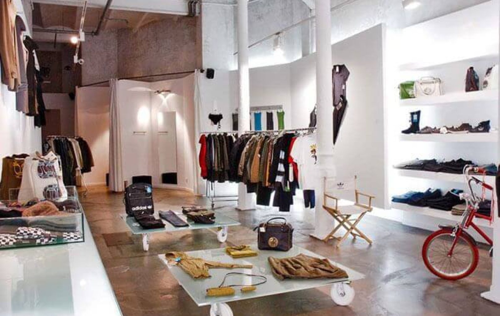 Clothing store interior in Barcelona with racks of clothes, shelves of bags and accessories, a red bicycle, and a table displaying folded garments on a polished concrete floor—just steps from the historic Montjuïc Castle.
