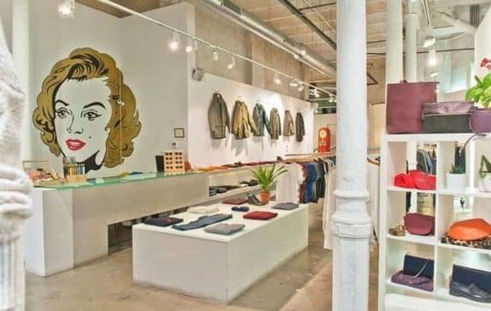 Moderna boutique IGUAPOP SHOP con ropa doblada en mesas de exposición, bolsos en estanterías, chaquetas en percheros de pared y un gran rostro de mujer ilustrado en la pared.
