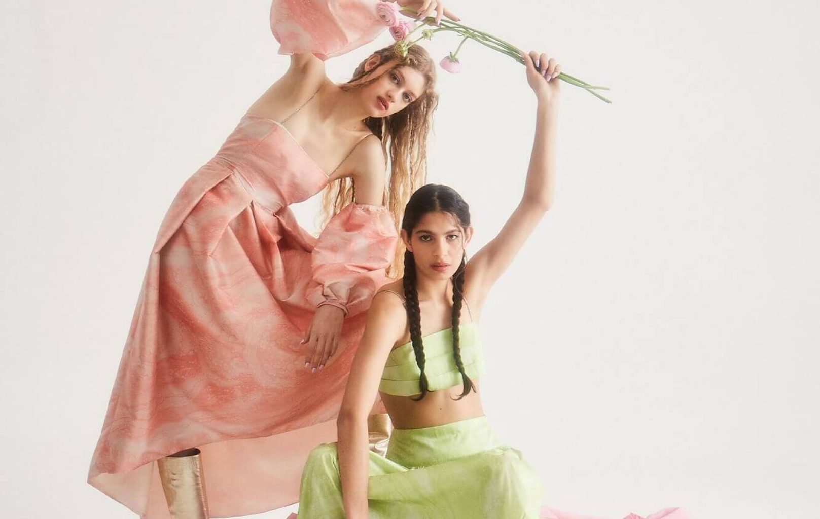 Two women in pastel dresses pose against a white background, evoking the elegance of Barcelona. One stands in a pink dress holding a flower, while the other sits in green with pink fabric draped around her, reminiscent of scenes near Montjuïc Castle.