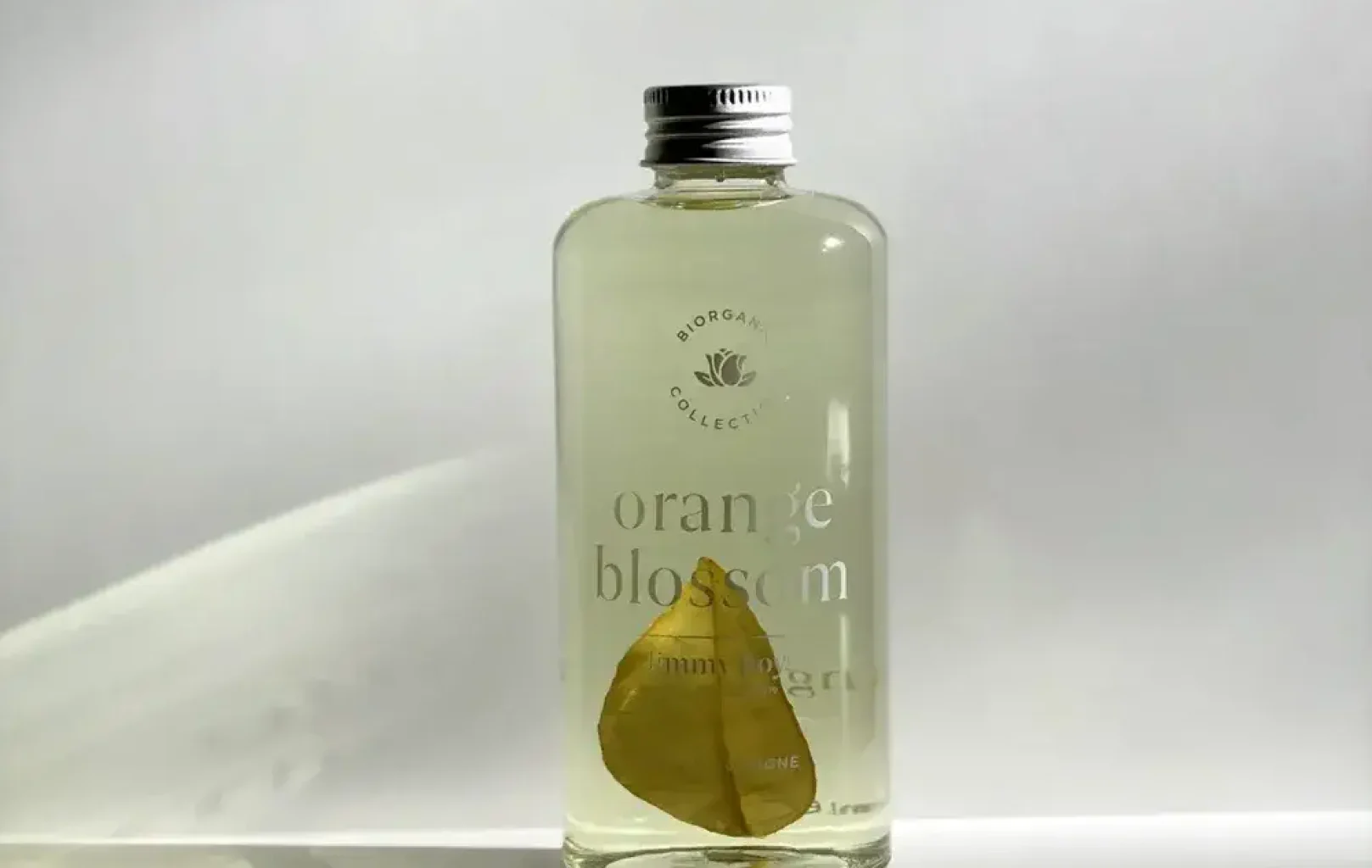 Clear glass bottle labeled "orange blossom," part of Jimmy Boyd fragrances, containing a light liquid and a single dried leaf, with a silver cap, set against a plain white background.