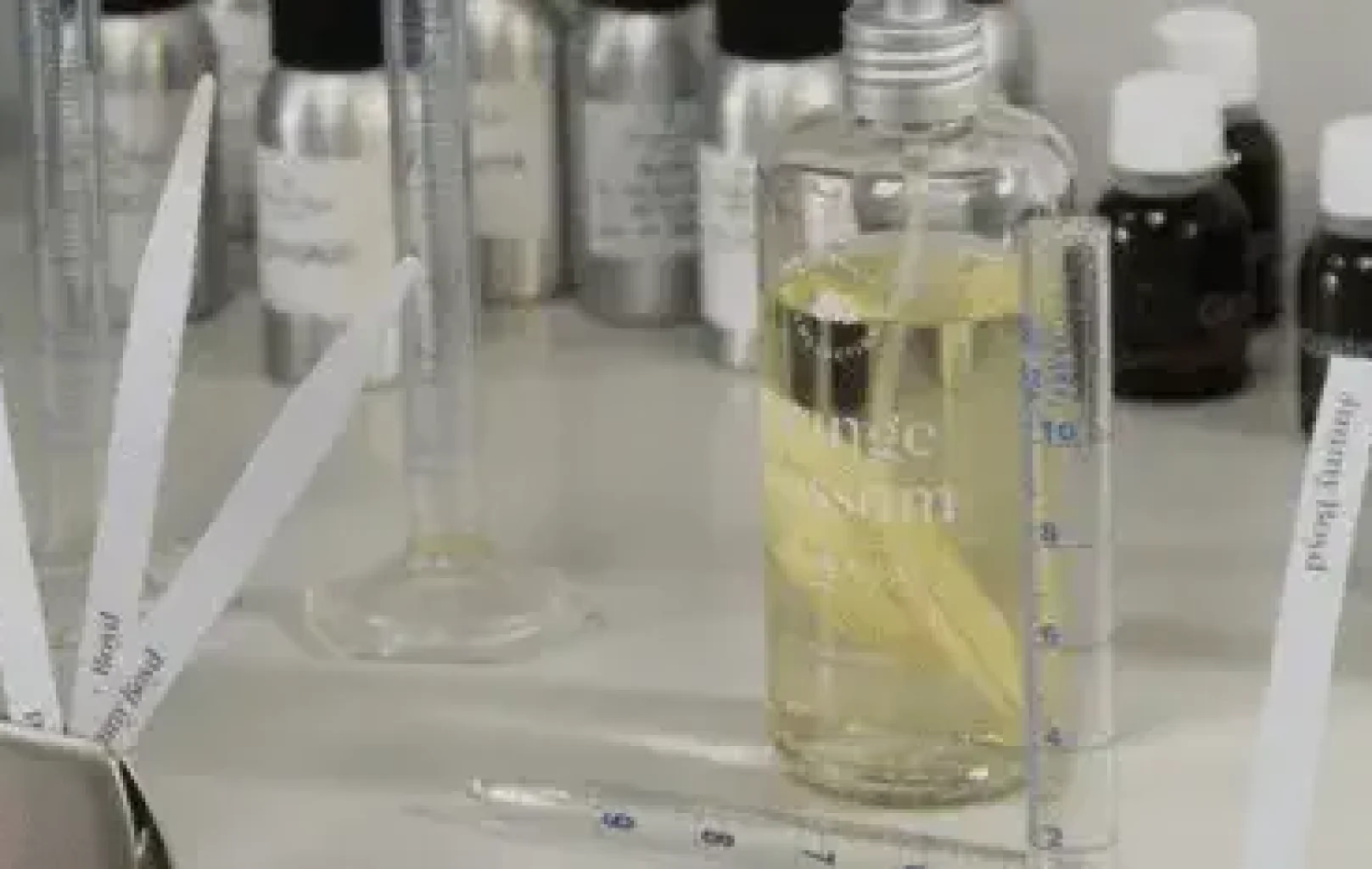 A clear bottle with yellow liquid, glass droppers, labeled metal and brown bottles, scent strips, and graduated cylinders on a laboratory workstation showcase the art of creating fragrances inspired by Jimmy Boyd.