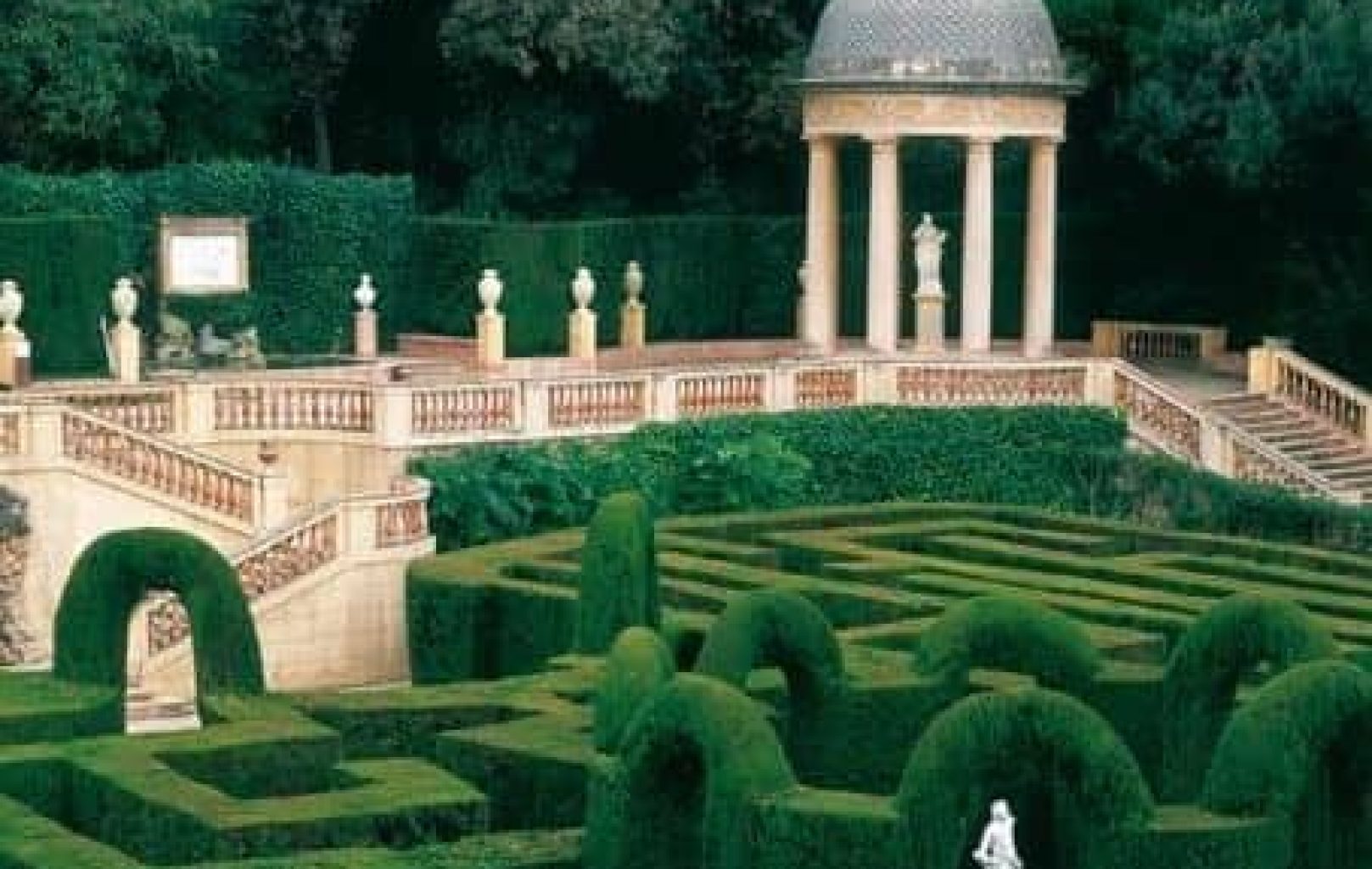 A formal garden inspired by the Horta Labyrinth in Barcelona park features a large hedge maze in the foreground and a domed pavilion with columns and statues in the background, all surrounded by lush greenery.
