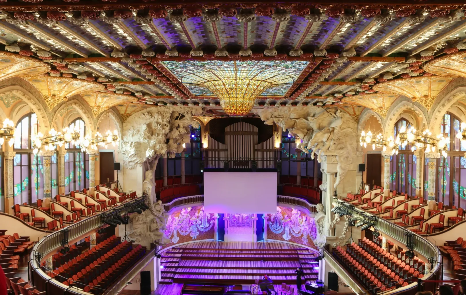Interior view of an ornate concert hall at L’Auditori Barcelona, featuring a stained glass ceiling, sculpted walls, chandeliers, red seats, a stage, and a large screen at the center.