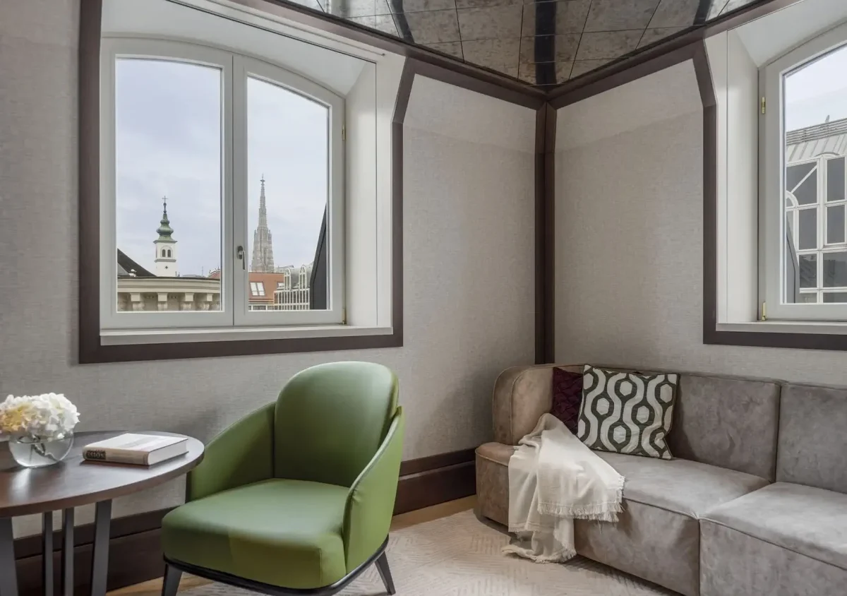 Modern lounge area with green chair, round table, and L-shaped sofa near windows overlooking a cityscape with church spires, exuding the refined elegance of a Palais Suite.