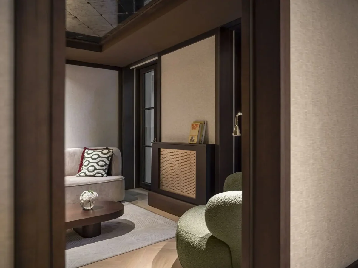 A modern family two bedroom living room featuring a beige sofa, patterned cushion, green armchair, wooden coffee table, and books on a cabinet shelf beside a window.