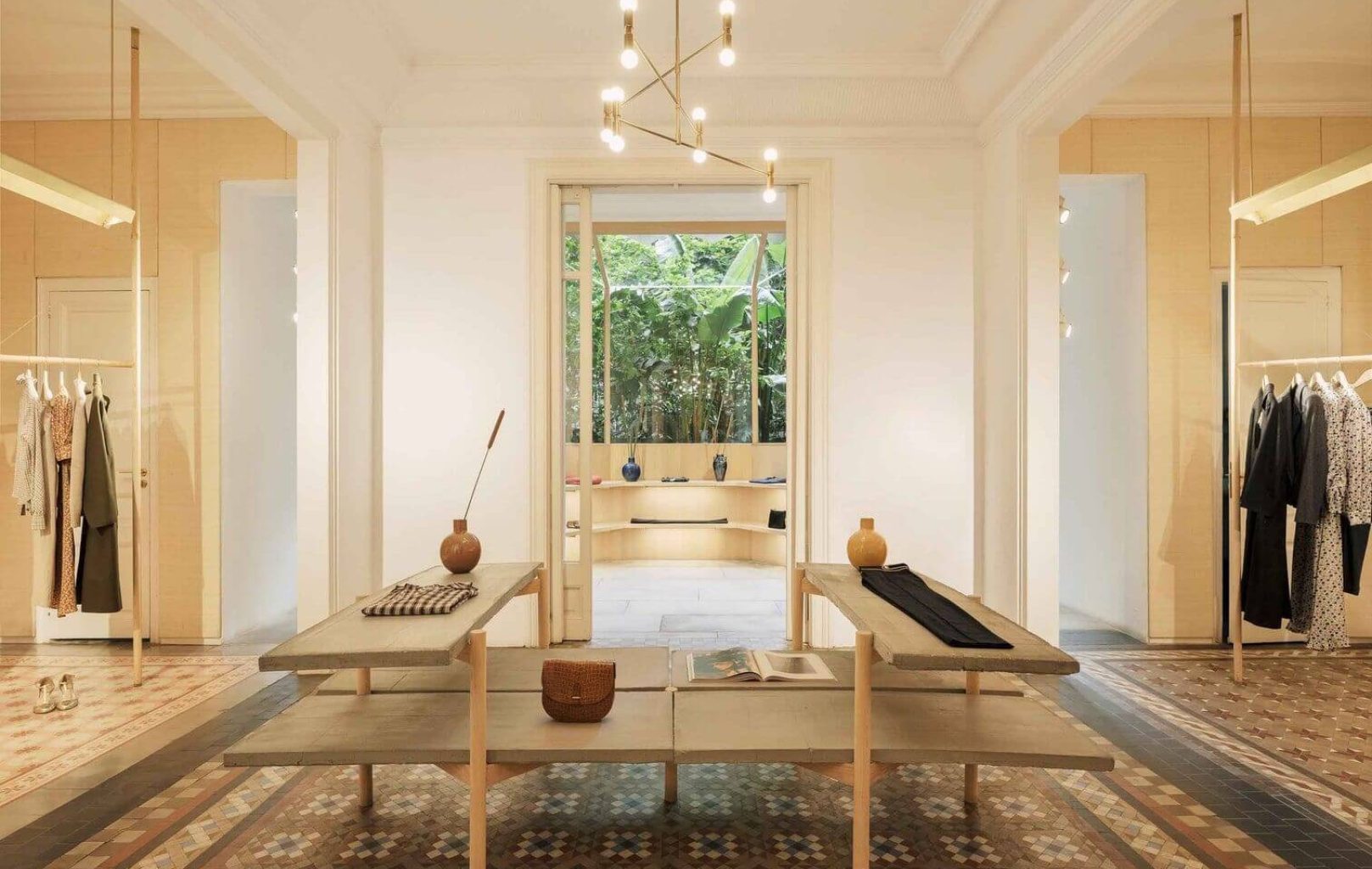 Interior de boutique minimalista con suelo de baldosas estampadas, dos mesas de exposición, lámparas colgantes, percheros a ambos lados y un gran ventanal que deja entrever una vegetación que recuerda a los jardines del castillo de Montjuïc de Barcelona.