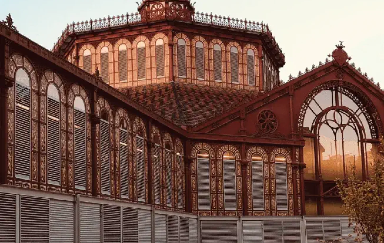 Un edificio histórico de Barcelona con herrajes ornamentados, ventanas arqueadas y una detallada decoración en la azotea recibe la cálida luz del sol, invitándole a saborear la rica arquitectura de la Tierra.