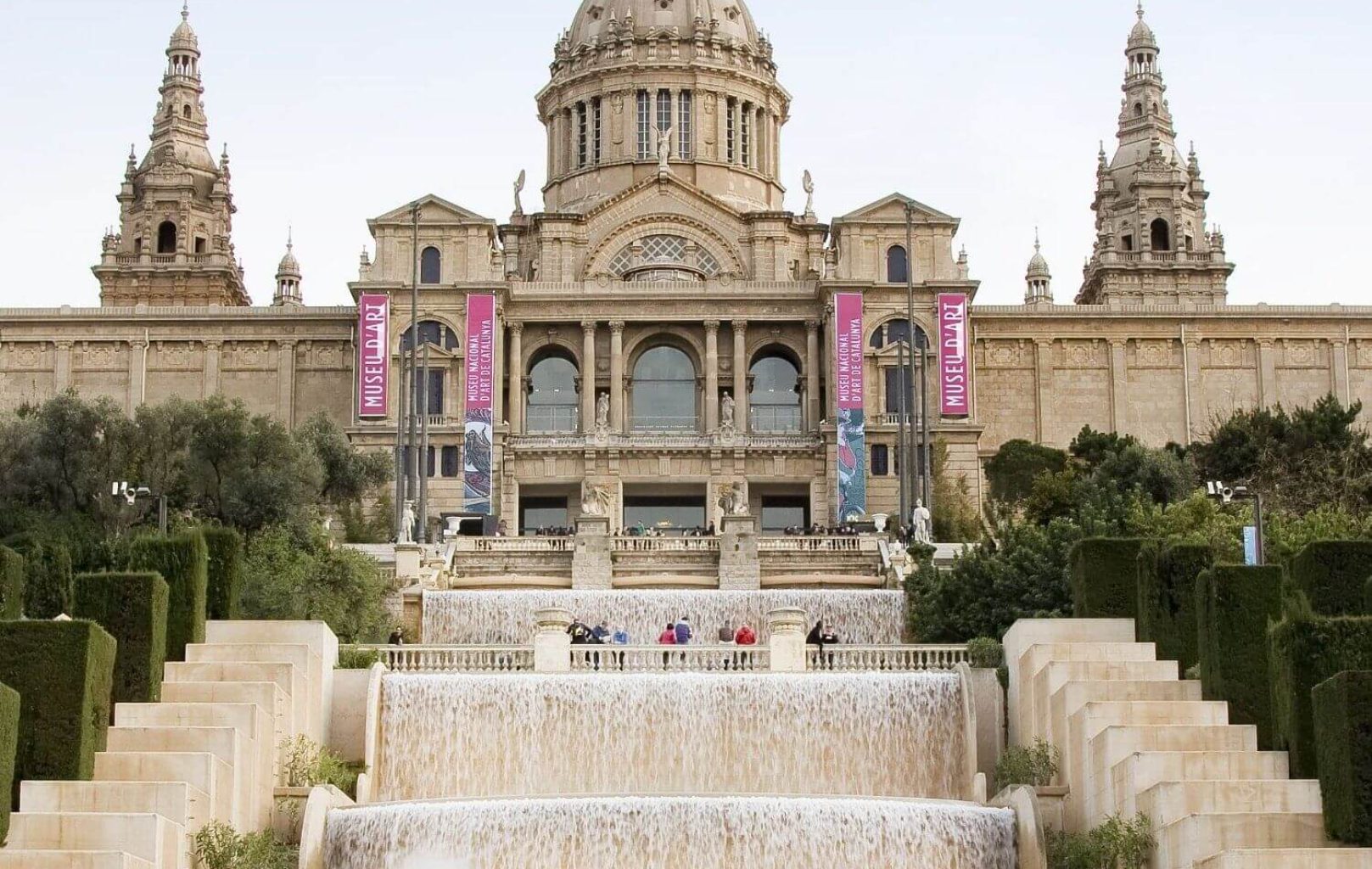 The image shows the Museu Nacional d'Art de Catalunya (MNAC) in Barcelona, with people walking near cascading fountains and the grand art museum building in the background.