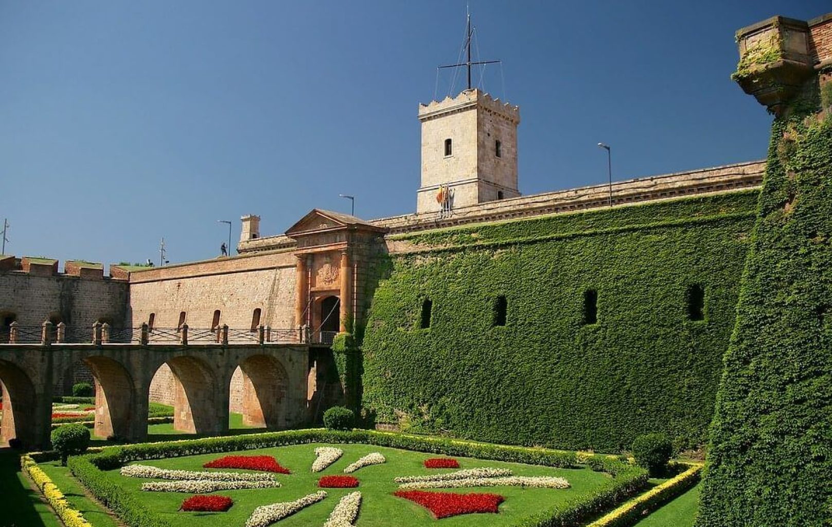 Montjuïc Castle, a historic fortress in Barcelona, boasts ivy-covered stone walls, an arched bridge, and a manicured garden with red and white flowerbeds beneath a clear blue sky.