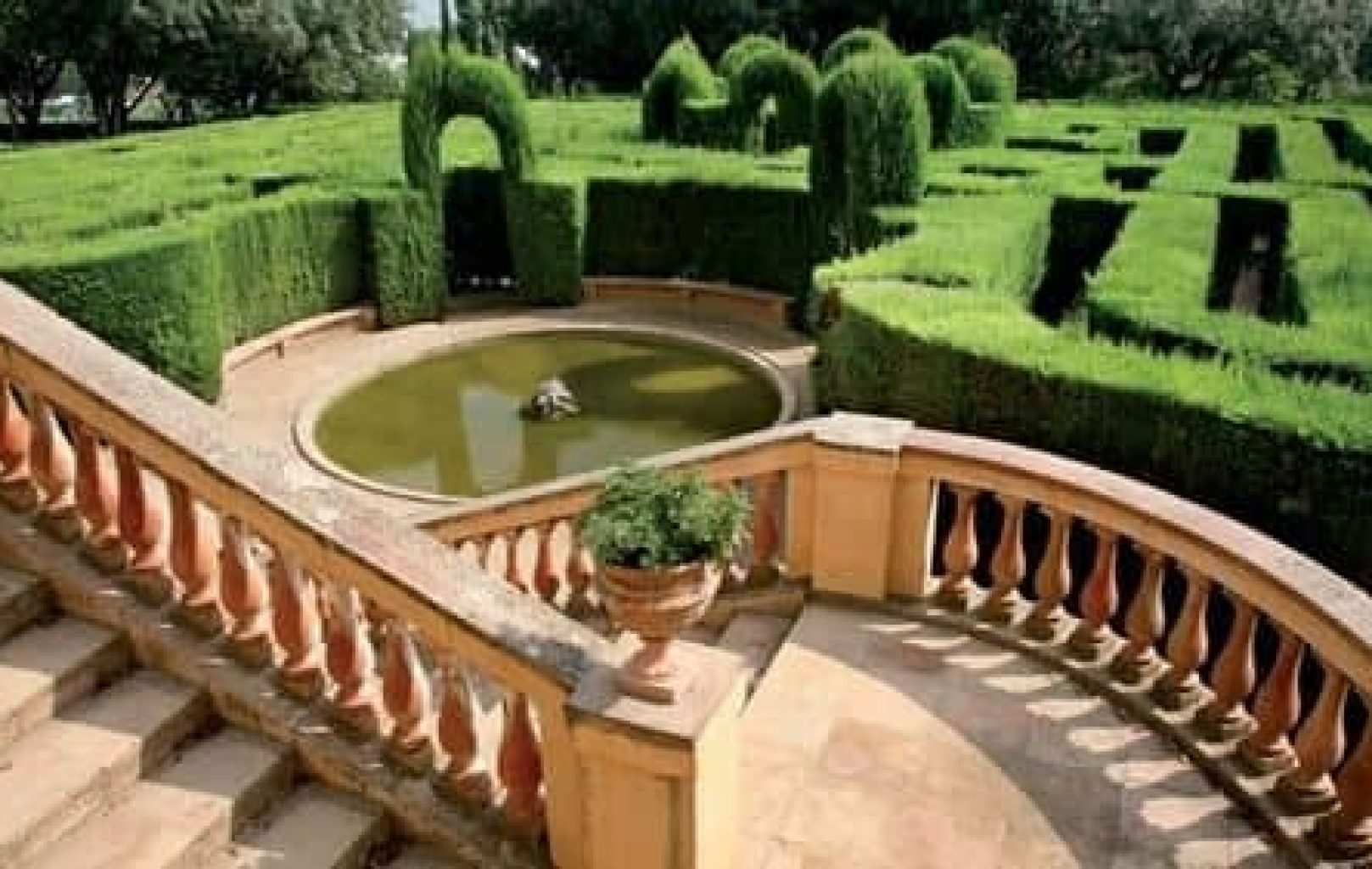 Stone staircase with balustrade leading to a circular pond, surrounded by manicured green hedges forming a labyrinth in the formal Parque Laberinto de Horta garden in Barcelona.