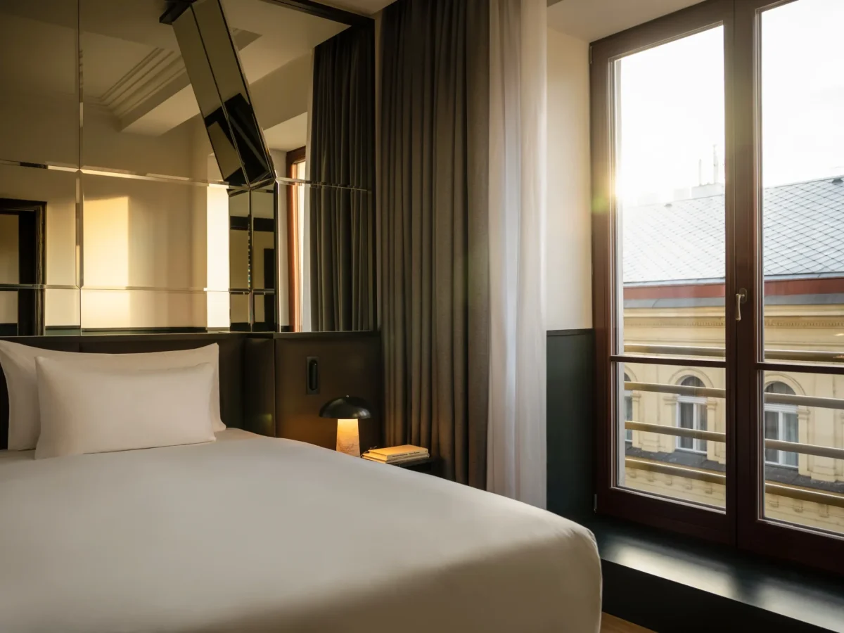 Moderne Executive Suite mit sauber gemachtem Bett, verspiegelter Wand, Nachttischlampe und großem Fenster mit Blick auf ein Gebäude bei Sonnenuntergang.