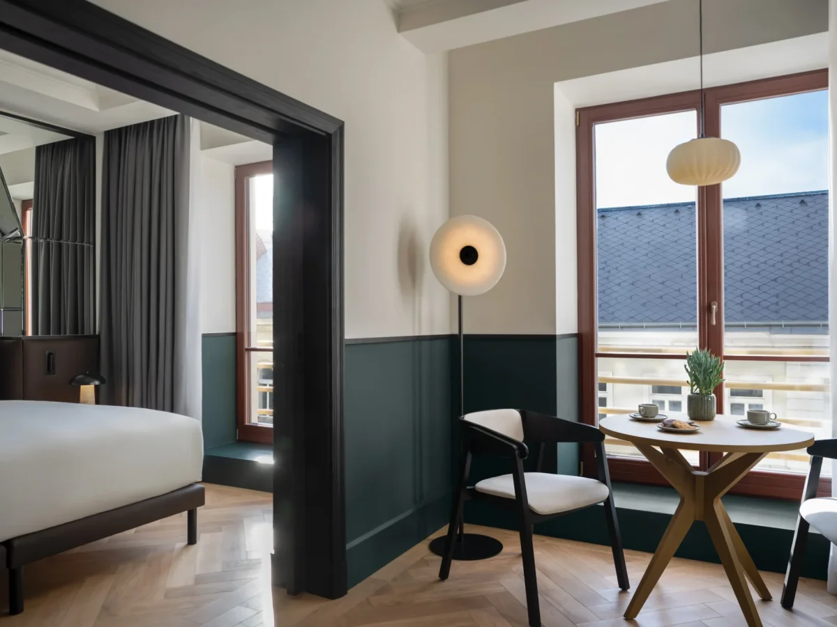 Modern Executive Suite featuring a bed, round table set for two, two chairs, large windows, and contemporary lighting. Natural light fills the space.