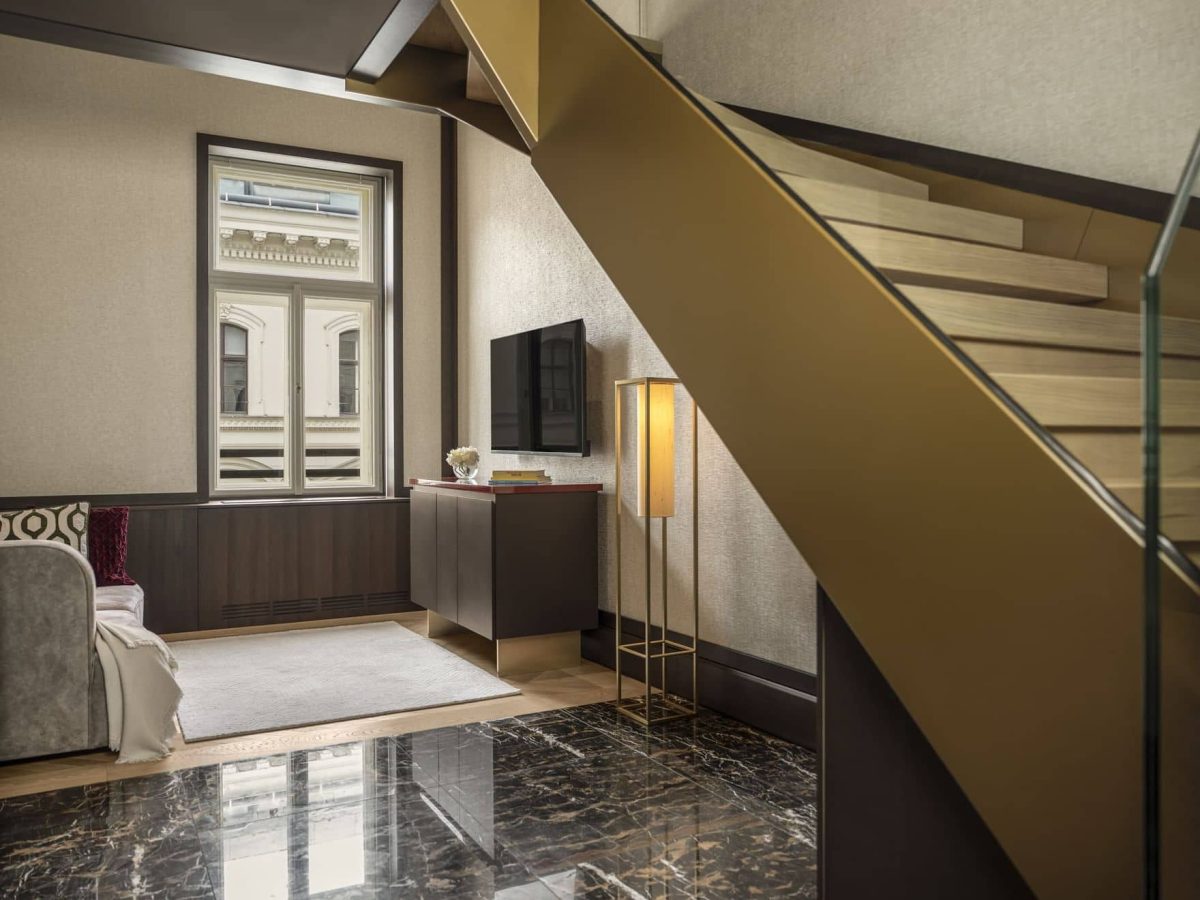 Modern living area with luxury flooring, beige walls, a wooden staircase, floor lamp, flat-screen TV, and a window showcasing an exterior building facade.