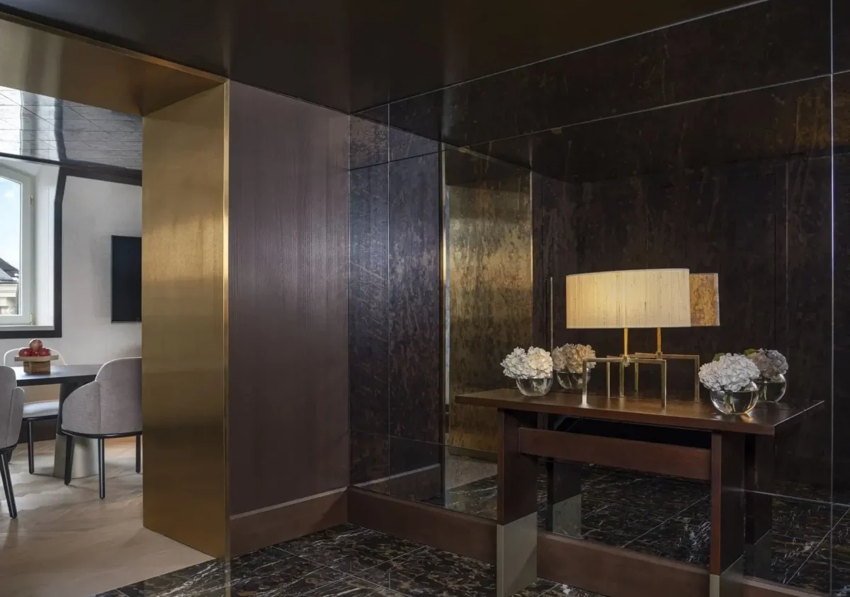 A modern interior in the Palais Suite features dark marble floors, a mirrored wall, and a console table with a lamp and vases of white flowers. Through an open doorway, a dining area with light chairs is visible.