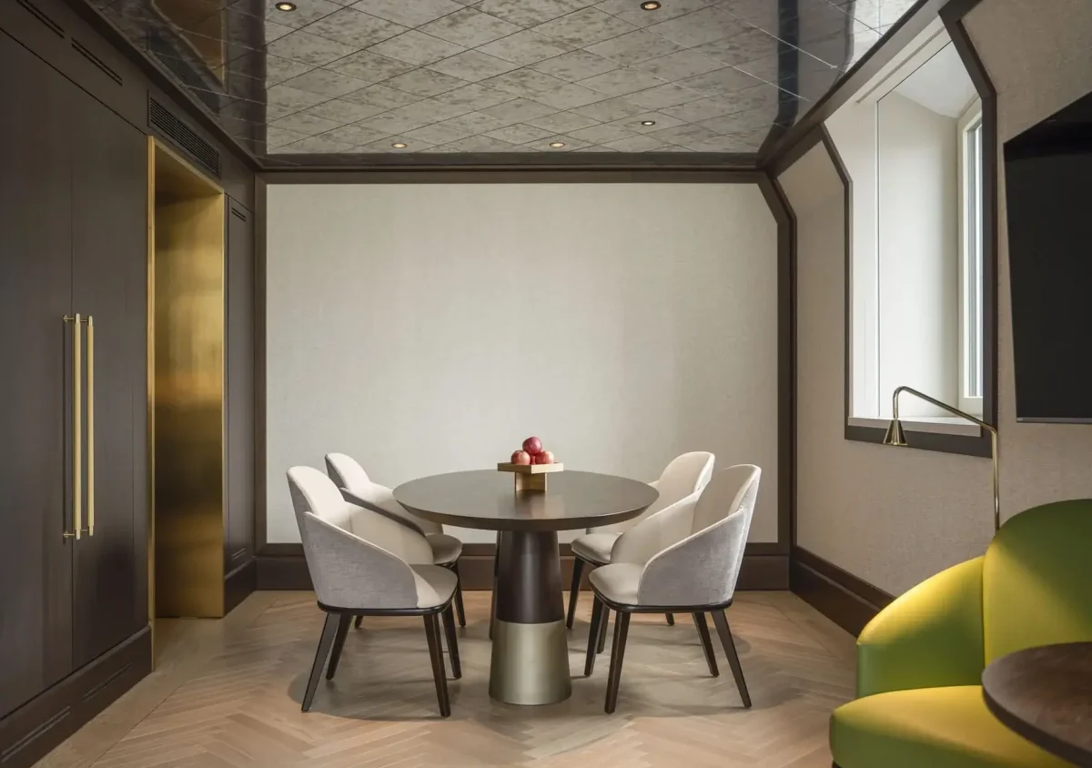 A modern meeting room in the Palais Suite features a round table, five upholstered chairs, a bowl of apples, wooden walls, large windows, and a green sofa in the corner.