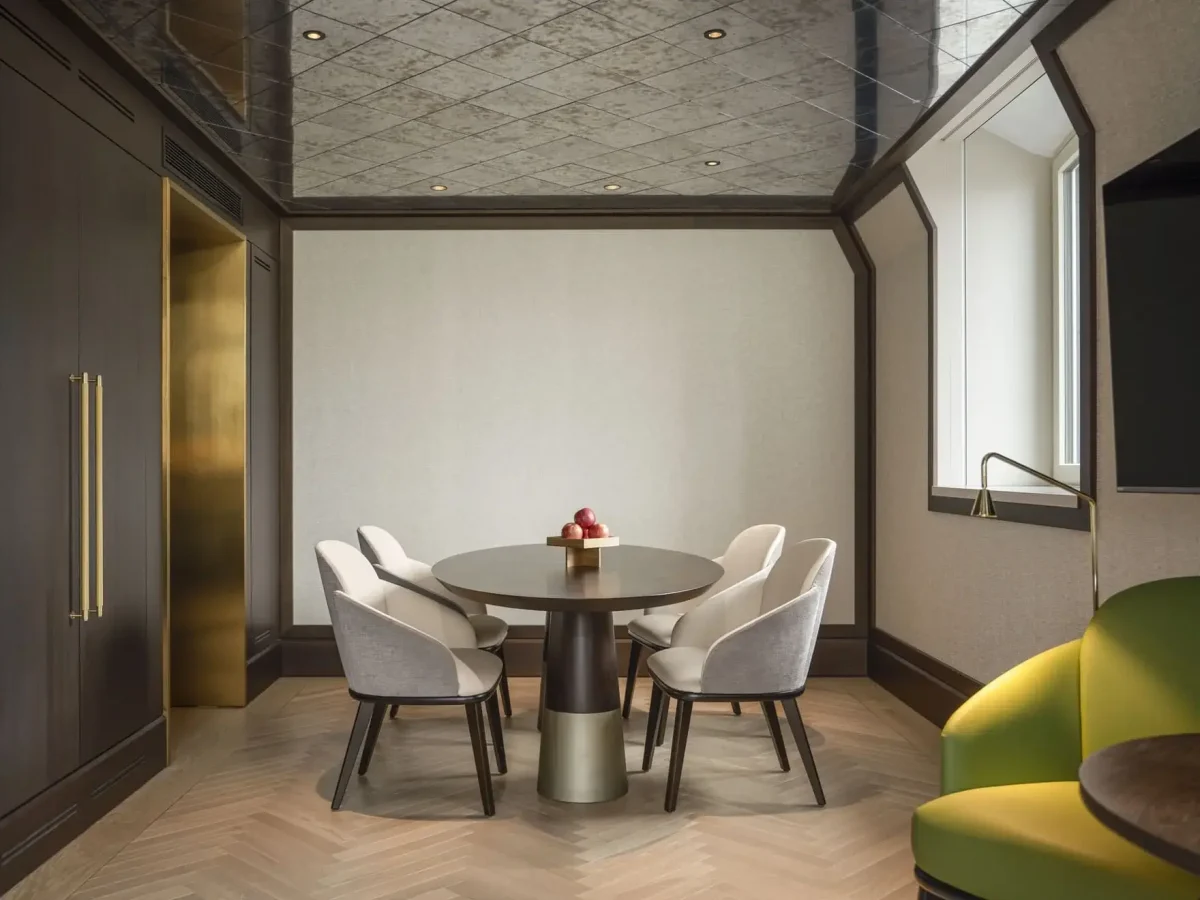 A modern meeting room in the Palais Suite features a round table, five upholstered chairs, a bowl of apples, wooden walls, large windows, and a green sofa in the corner.