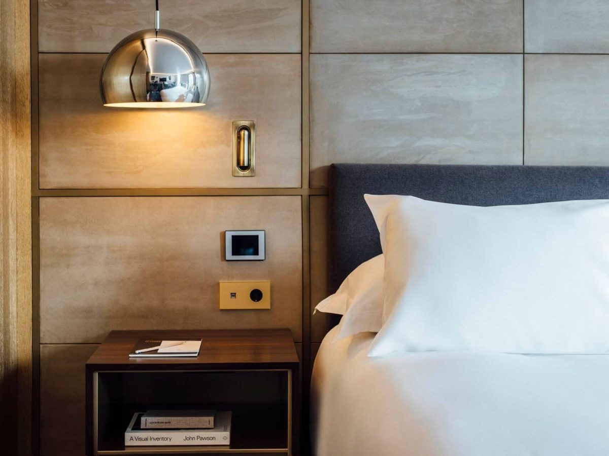 En la Sala Galería hay una cama bien hecha con sábanas blancas, junto a una mesilla de noche de madera, una lámpara moderna, interruptores de pared y libros en una estantería.