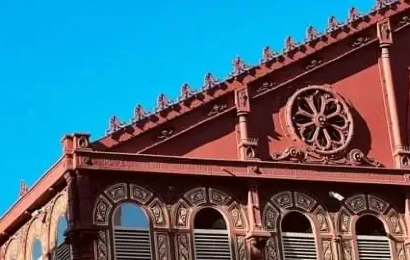 Fachada roja ornamentada de un edificio histórico de Barcelona con ventanas arqueadas, molduras decorativas y un detalle de ventana circular bajo un cielo azul despejado.