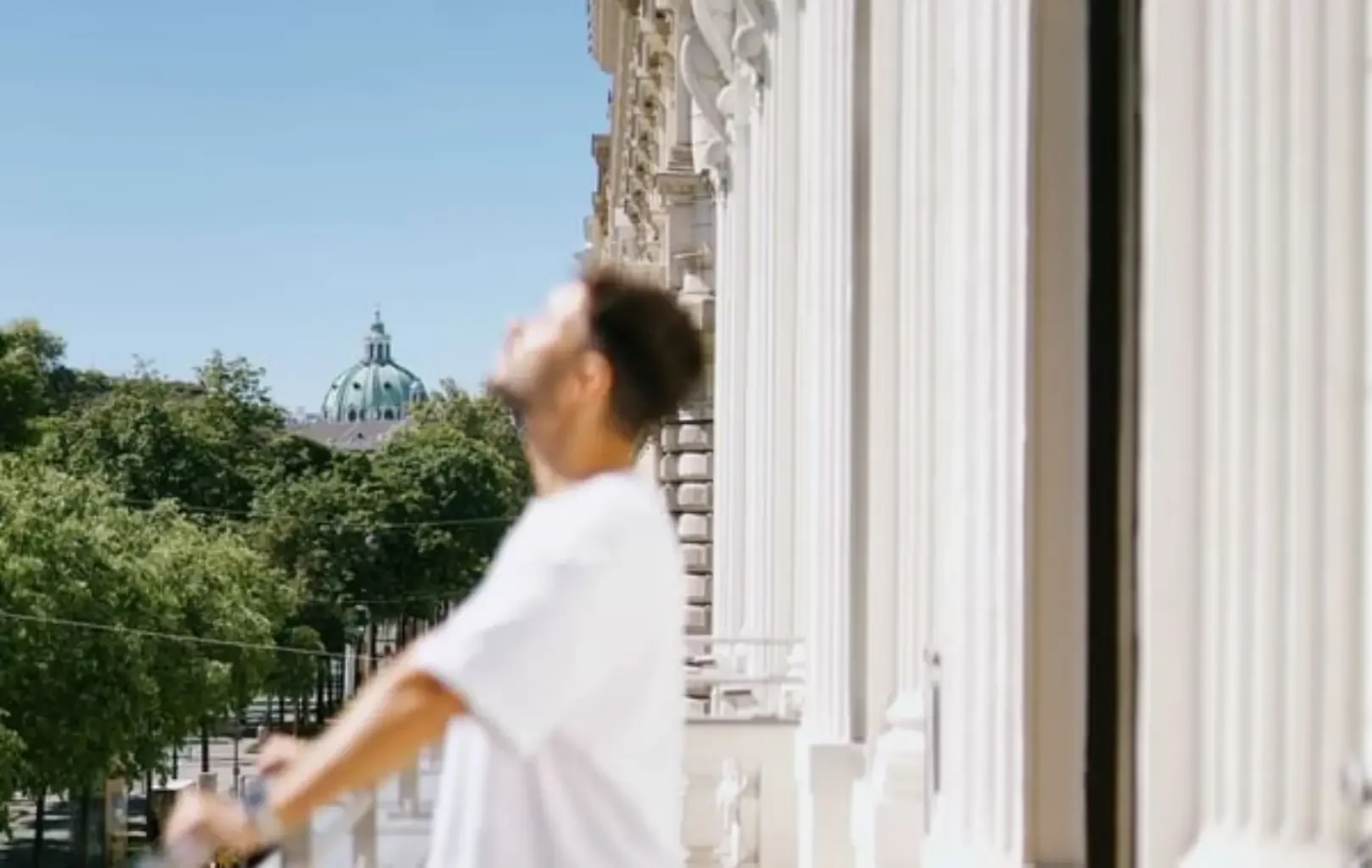 Eine Person in einem weißen Hemd steht auf einem Balkon mit Säulen und genießt eine Suite Erlebnis, während sie nach oben blickt, mit Bäumen und einem Kuppelgebäude im Hintergrund.