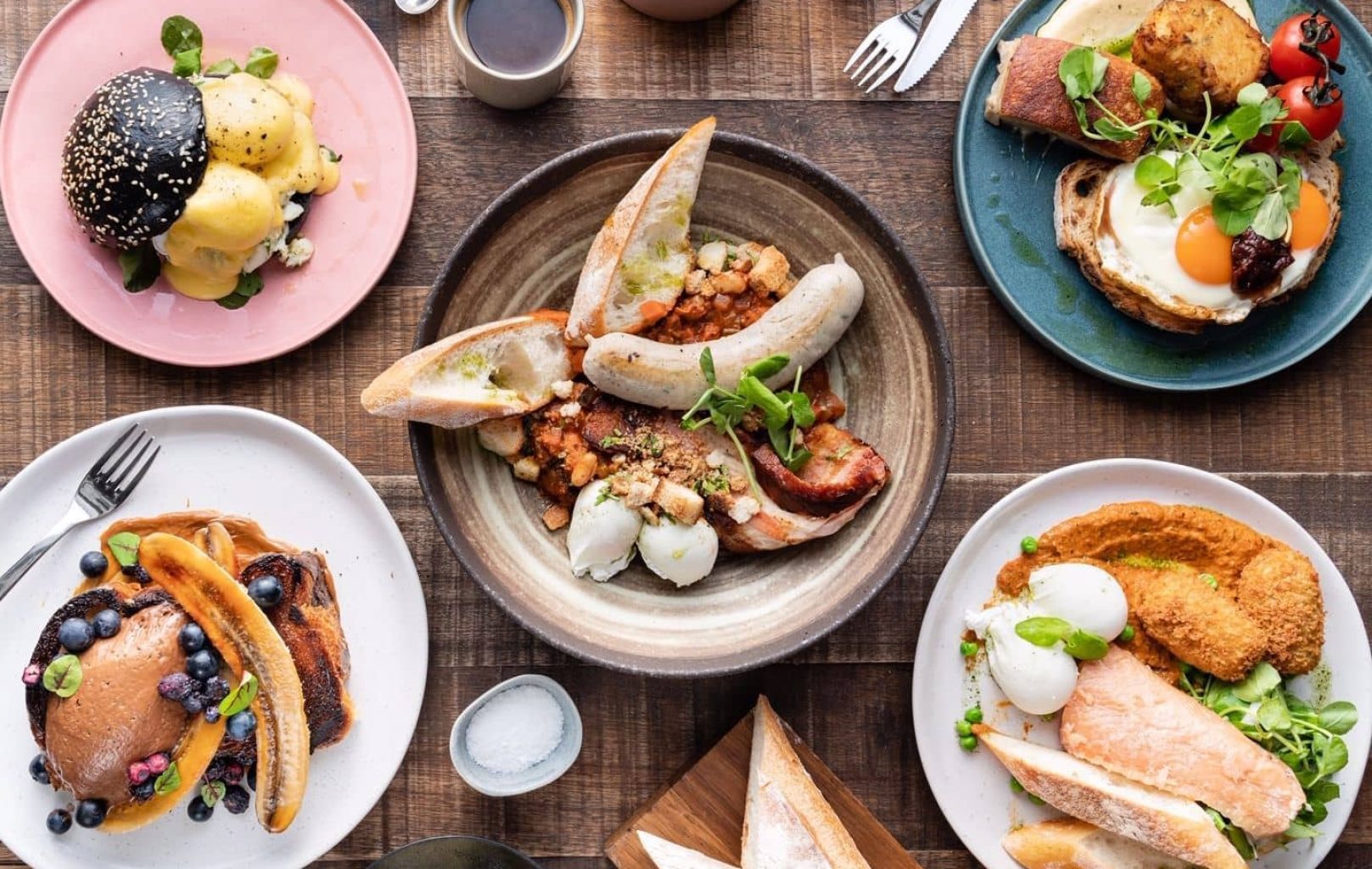 A table set with assorted breakfast dishes, including eggs, sausages, toast, and pancakes with blueberries at Hardware Société Barcelona, alongside grilled vegetables and coffee on a wooden surface.