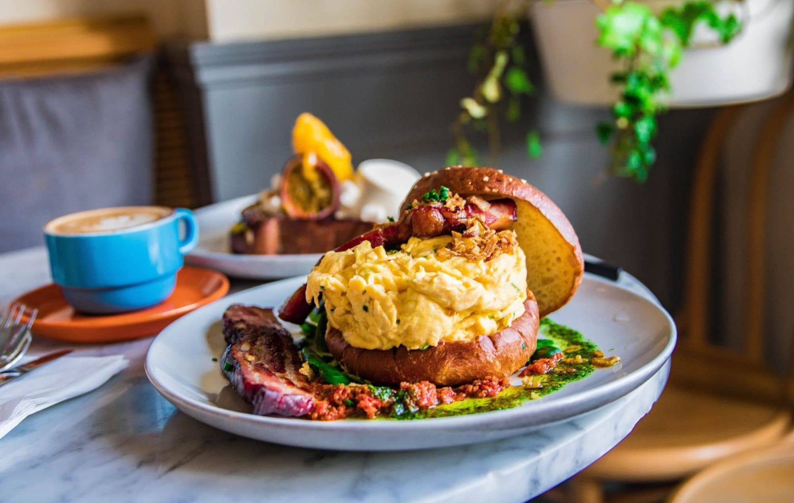 A breakfast sandwich with scrambled eggs and bacon on a bun is served on a white plate with sauce and a vegetable at Hardware Société Barcelona; a coffee cup and another plate are in the background.