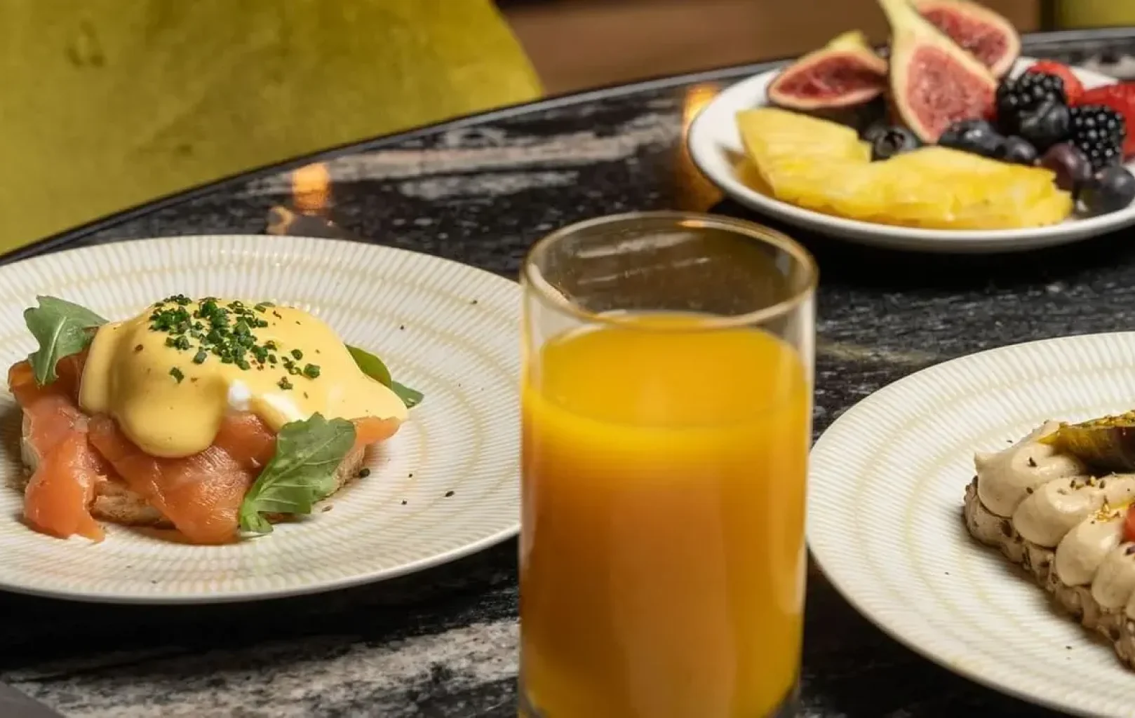 A glass of orange juice, smoked salmon eggs benedict, toast with cream spread, a plate of assorted fruit, and a dessert are arranged on a marble table—perfect for an Autumn Escape brunch.
