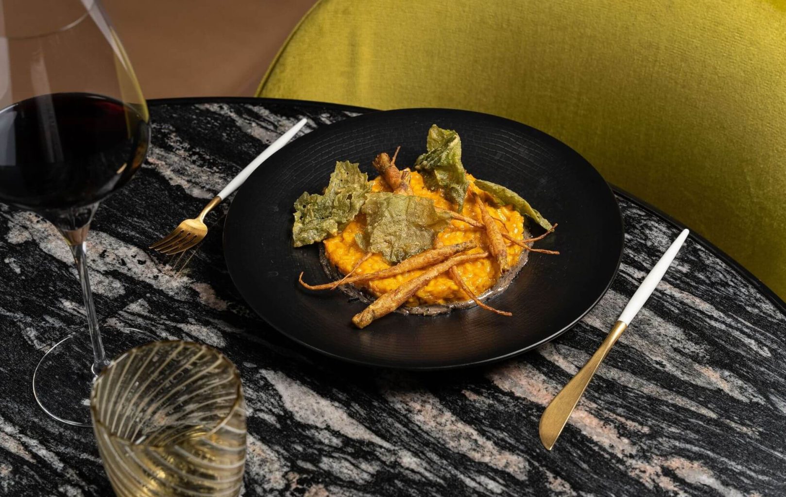 En una mesa de mármol negro, junto a una copa de vino tinto y un vaso de agua, hay un plato de risotto cremoso con verduras y tubérculos fritos.