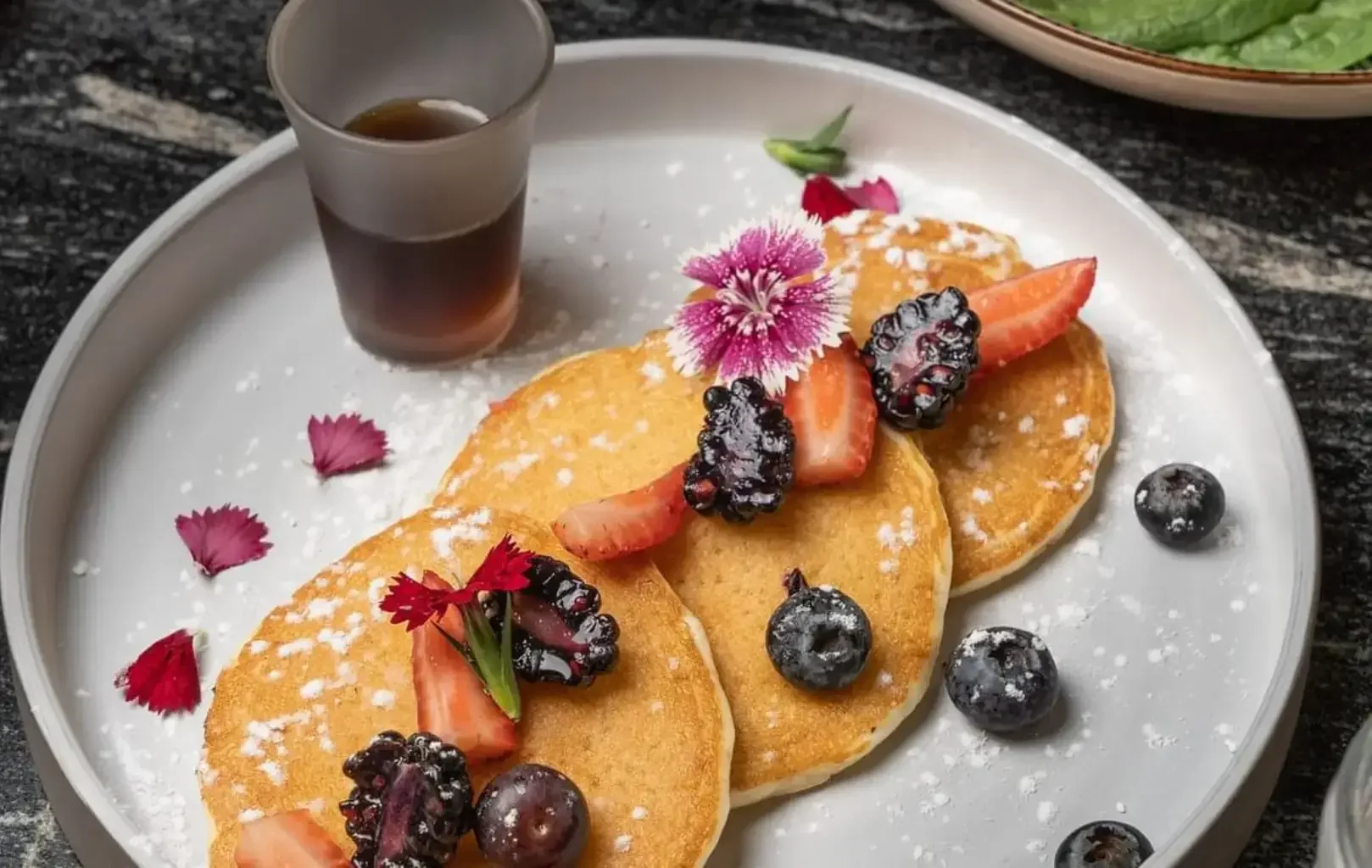 Tres creps de Benestar, cobertes amb maduixes, móres, nabius i flors comestibles a rodanxes, servides amb una tassa de xarop en un plat blanc.