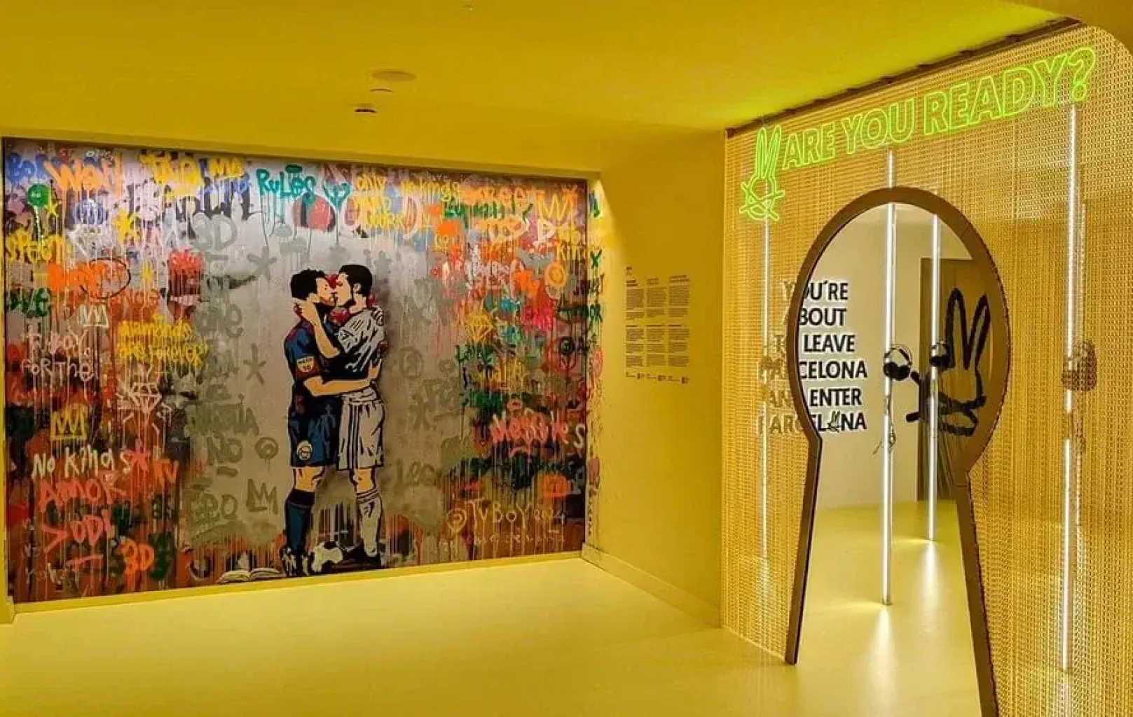 Un pasillo amarillo brillantemente iluminado del Museo del Conejo Blanco presenta un colorido mural de graffiti de dos jugadores de fútbol besándose y una puerta con un letrero de neón en el que se lee "ARE YOU READY?" (¿estás listo?) y un texto en el interior de la puerta.