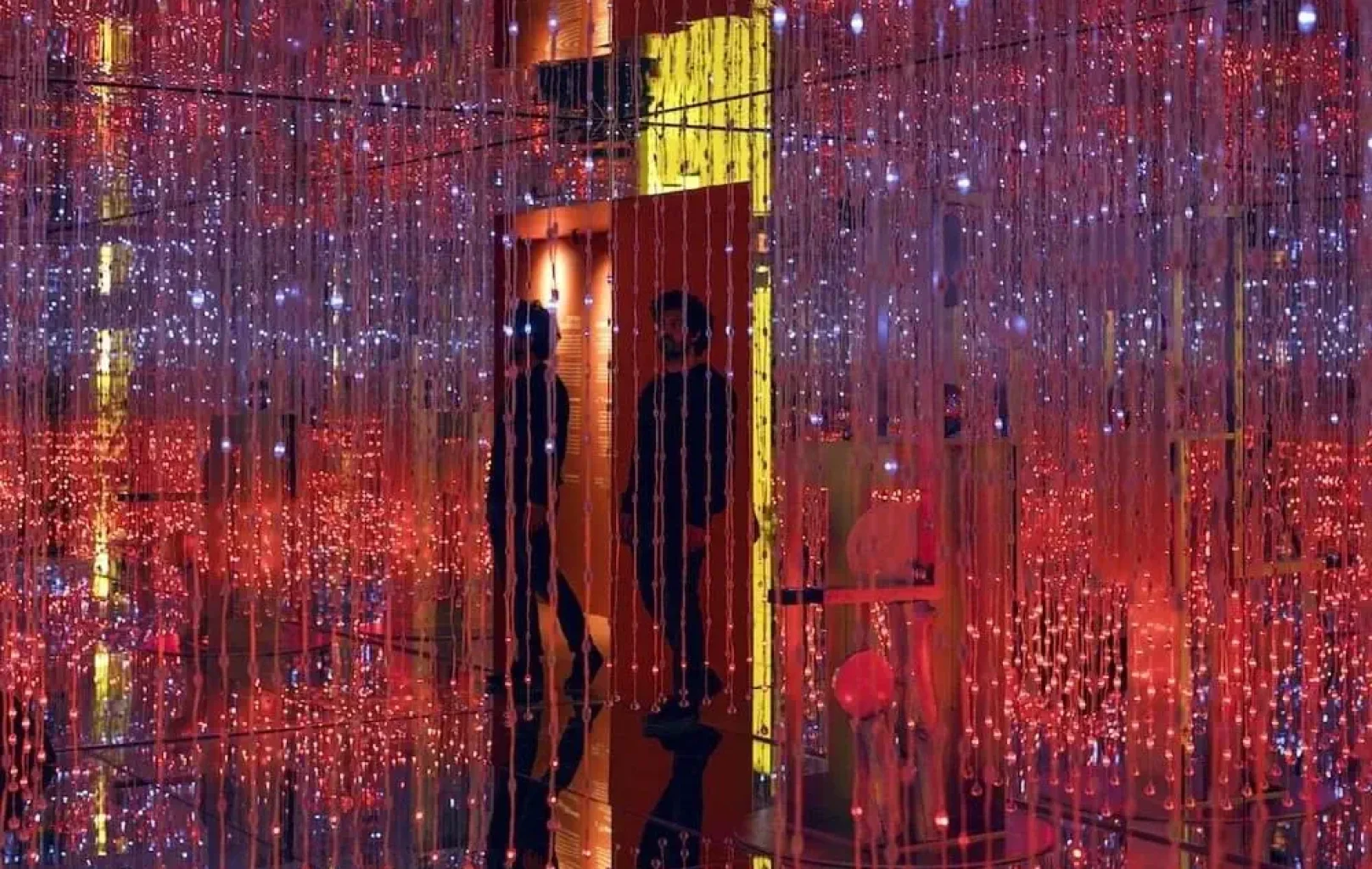 Dos personas permanecen de pie en una sala del Museo White Rabbit, rodeadas de hilos colgantes de luces LED rojas y blancas que crean una instalación luminosa vibrante y envolvente.
